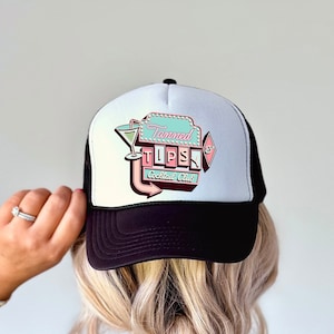 Op de afbeelding: Zwarte en witte truckercap met een roze, blauwe en groene logo met de tekst "Tanned Tops Cocktail Club".