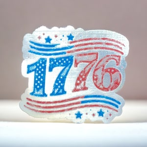 Puede incluir: Una pegatina plateada con un diseño de bandera estadounidense roja, blanca y azul y el número 1776 en azul.