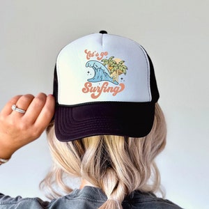 Könnte beinhalten: Eine schwarz-weiße Trucker-Kappe mit einem weißen Frontpanel, das eine farbenfrohe Illustration einer Welle, Palmen und den Text "Let's go Surfing" zeigt.