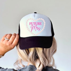 May include: A black and white trucker hat with a white mesh back. The front of the hat features a pink heart with a pearl border and the text "FUTURE Mrs." in pink.