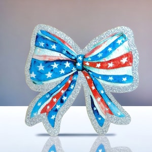 Puede incluir: Un lazo de purpurina plateado con un diseño de bandera estadounidense roja, blanca y azul.