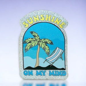 May include: A silver glitter iron-on patch with a blue background featuring a palm tree and a beach chair. The text "Sunshine on my mind" is written in yellow letters.