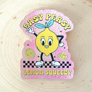 Könnte beinhalten: Ein rosa und gelber Aufkleber mit einer Cartoon-Zitronenfigur. Die Zitrone trägt ein Lächeln und hat große Augen. Der Aufkleber lautet "Easy Peasy Lemon Squeezy" in einer Retro-Schrift.