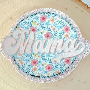 Könnte beinhalten: Ein weißer und blauer Kreis mit floralem Muster und dem Wort "Mama" in rosa-weißer Schrift. Der Kreis hat einen glitzernden Rand.