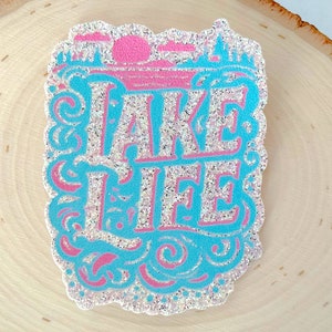 May include: A glittery sticker with the words "TAKE LIFE" in a stylized font. The design features a pink sun and clouds, with a turquoise border and lettering. The sticker is on a wooden surface.