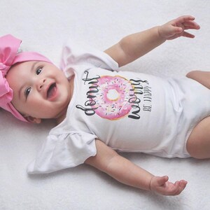 May include: A baby wearing a white onesie with a pink donut graphic and the text "Donut Worry Be Happy". The baby is wearing a pink bow headband and is lying on a white surface.