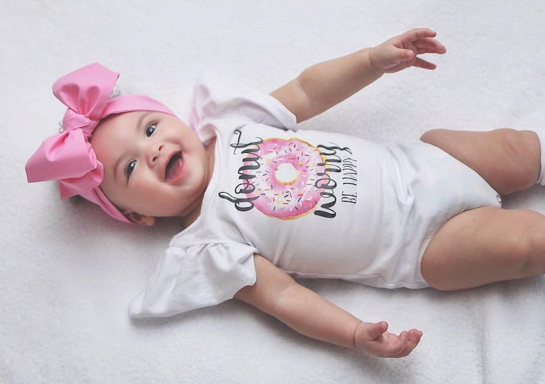 May include: A baby wearing a white onesie with a pink donut graphic and the text "Donut Worry Be Happy". The baby is wearing a pink bow headband and is lying on a white surface.