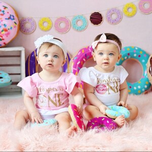May include: Two babies wearing pink and white donut-themed onesies with the text "Harper is One" and "Kinsley is One". The babies are sitting on a fluffy pink rug with donut-shaped toys.