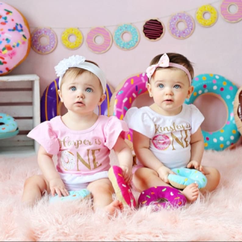 May include: Two babies wearing pink and white donut-themed onesies with the text "Harper is One" and "Kinsley is One". The babies are sitting on a fluffy pink rug with donut-shaped toys.