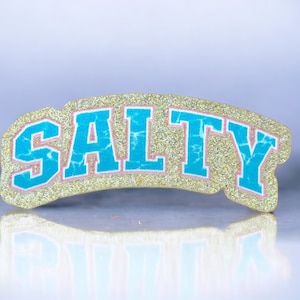 May include: A gold glitter sticker with the word "SALTY" in blue letters with a pink outline. The sticker is shaped like an arch.