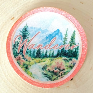 Può includere: Un adesivo rotondo con bordo rosa con un'illustrazione ad acquerello di una catena montuosa e una foresta. Il testo "Wanderer" è scritto in corsivo rosa sull'immagine.