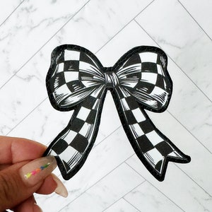 May include: A black and white checkered bow with a black border. The bow is held up against a white and gray marble background. The bow is a decorative accessory.