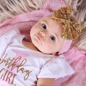 May include: A baby wearing a white onesie with the text "1st Birthday Girl" in pink and gold glitter. The baby is wearing a gold glitter headband with a pink bow.