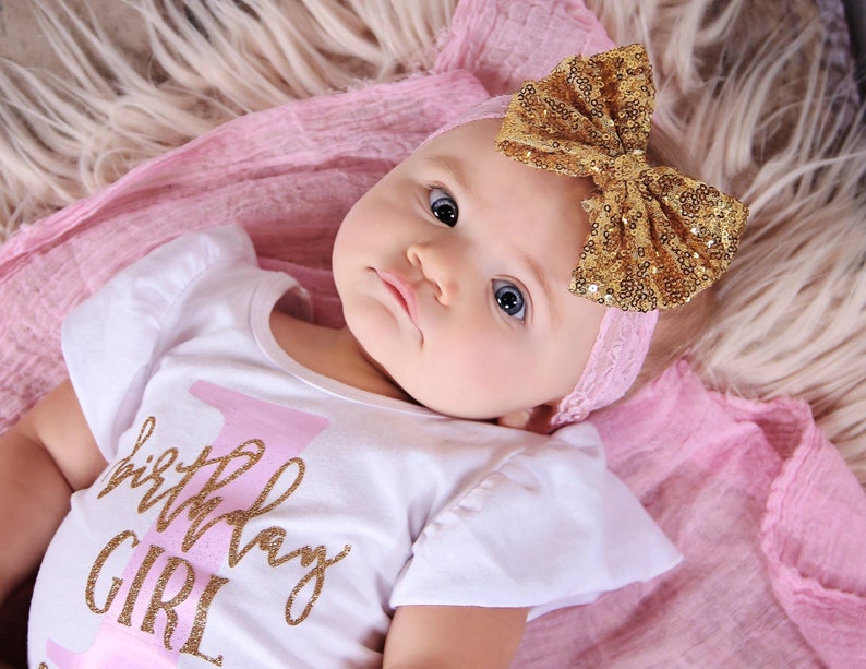 May include: A baby wearing a white onesie with the text "1st Birthday Girl" in pink and gold glitter. The baby is wearing a gold glitter headband with a pink bow.