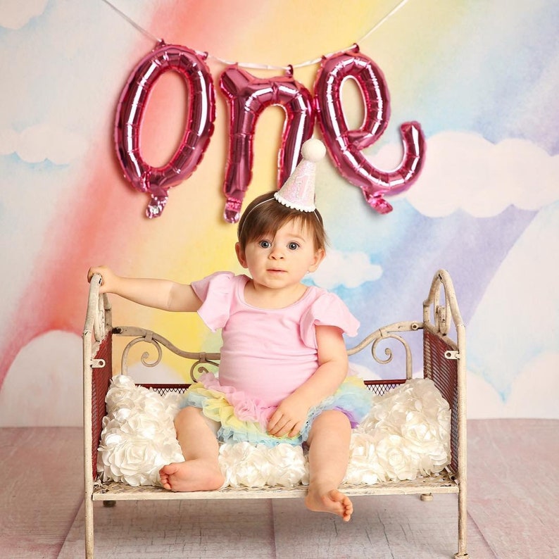 May include: A baby girl wearing a pink shirt and a rainbow tutu sits on a white metal bed with a pink "one" balloon banner behind her.