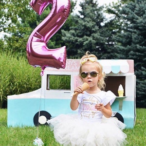 May include: A young girl wearing a white tutu skirt and a white t-shirt with the text "gwendolyn is two sweet" and a pink ice cream cone graphic. She is holding a waffle cone ice cream and wearing sunglasses. A large pink number 2 balloon is in the background.