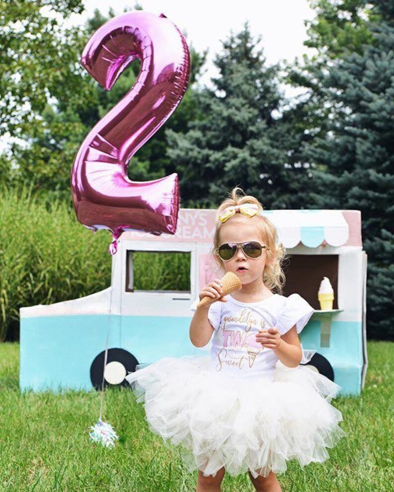 May include: A young girl wearing a white tutu skirt and a white t-shirt with the text "gwendolyn is two sweet" and a pink ice cream cone graphic. She is holding a waffle cone ice cream and wearing sunglasses. A large pink number 2 balloon is in the background.