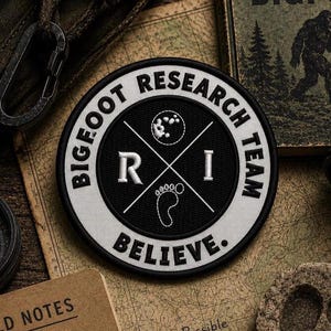 May include: A circular embroidered patch with a black center and a white border. The text reads BIGFOOT RESEARCH TEAM BELIEVE. The center features a white cross, the letters R and I, a moon icon, and a footprint icon in white thread.