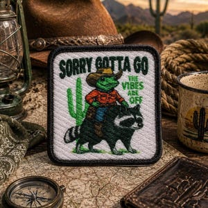 May include: A square embroidered patch featuring a green frog wearing a brown cowboy hat and orange shirt, riding a black and white raccoon. The text reads SORRY GOTTA GO and THE VIBES ARE OFF, with a green cactus graphic on a white background.