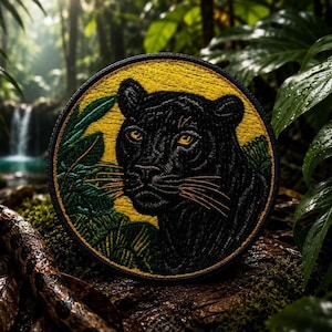 May include: Round embroidered patch featuring a black panther with yellow eyes and whiskers, set against a yellow background. Green leaves and a black border complete the design. The patch is likely for clothing or accessories.