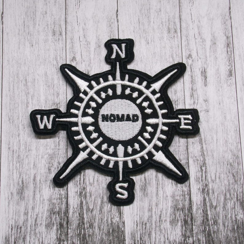 May include: A black and white embroidered patch featuring a compass rose design. The center circle contains the word NOMAD in black text. Four cardinal directions are marked with the letters N, S, E, and W at the ends of the compass points.