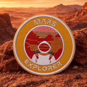 May include: A circular embroidered patch featuring a yellow border with the white text MARS EXPLORER. The center depicts a white astronaut figure against a red and orange landscape with a pink sky, all rendered in detailed thread work.