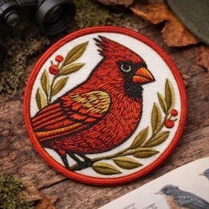 Peut inclure: Un écusson brodé circulaire représentant un cardinal rouge vif avec un masque facial noir et un bec orange. L'oiseau est perché parmi des feuilles vert olive et de petites baies rouges, le tout sur un fond blanc avec une bordure rouge.