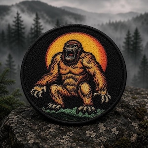 May include: Circular black patch with an orange-brown ape-like figure, mouth open, against a yellow and orange sun. The background shows a forest and mountains in a misty scene.