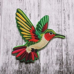 May include: A colorful embroidered patch of a hummingbird in flight. The hummingbird is green, red, yellow, and white with a long beak.