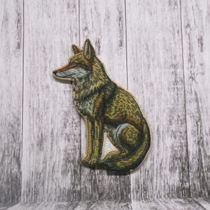 May include: An embroidered wolf, brown and grey in colour, sitting on a white wooden background.