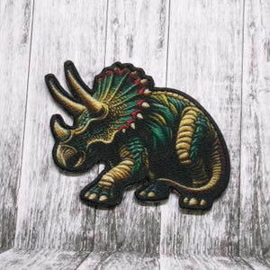 May include: A green, gold, and black embroidered patch of a triceratops dinosaur. The patch is detailed with a textured fabric and has a black border.