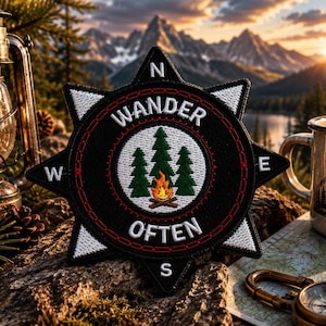 May include: A star-shaped embroidered patch featuring a black background with white cardinal directions N, E, S, and W. The center displays three green pine trees and a campfire with the white text WANDER OFTEN arched above and below the design.
