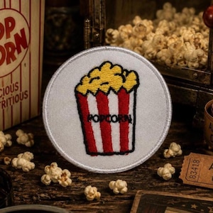 May include: A circular embroidered patch featuring a white background with a central graphic of a red and white striped popcorn container filled with yellow popcorn. The word POPCORN is stitched in black thread across the red and white stripes.