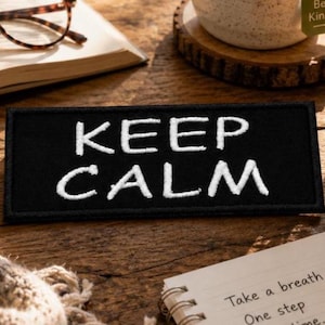 May include: A rectangular black embroidered patch featuring the white text KEEP CALM in a bold, sans-serif font. The patch has a clean, stitched border and is presented as a flat, rectangular fabric accessory with high-contrast lettering.
