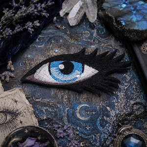 May include: An embroidered patch shaped like an eye with a bright blue iris, a black pupil, and white sclera. The eye is surrounded by thick, black stitched eyelashes extending to the right, creating a stylized, elongated almond shape.