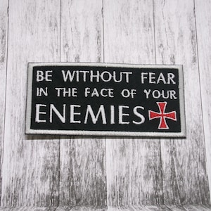 May include: Black rectangular patch with white embroidered text: "BE WITHOUT FEAR IN THE FACE OF YOUR ENEMIES." A red cross is on the right. The patch has a white border and is on a wood-grain background.