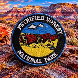May include: A circular embroidered patch featuring a blue sky, yellow ground, and red petrified wood logs. The black border contains the white text PETRIFIED FOREST NATIONAL PARK. The patch has a blue outer edge and a detailed stitched texture.