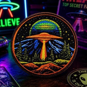 Pode incluir: Um emblema bordado circular apresentando um OVNI laranja vibrante pairando sobre uma cordilheira azul-escura e pinheiros verdes. Um grande planeta multicolorido situa-se no céu noturno estrelado acima da nave, com colinas alaranjadas.