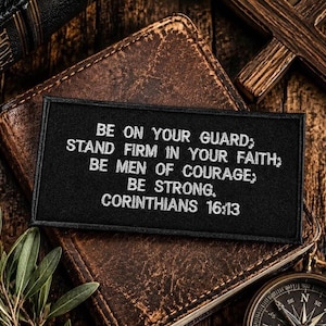 May include: Black patch with white embroidered text: "BE ON YOUR GUARD; STAND FIRM IN YOUR FAITH; BE MEN OF COURAGE; BE STRONG. CORINTHIANS 16:13." The patch rests on a leather-bound book, with a wooden cross and compass in the background.
