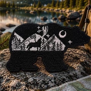 May include: A black embroidered patch shaped like a bear. The interior features a white stitched landscape of a howling wolf, pine trees, and mountains under a crescent moon and stars. The patch has a textured, thread-based surface finish.