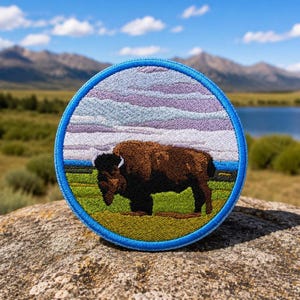 May include: Embroidered patch featuring a brown bison grazing on green grass under a purple and blue sky. The round patch has a blue border and depicts a mountain range and lake in the background. A nature-themed accessory.