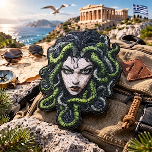 May include: Embroidered patch featuring Medusa with green snake hair and piercing green eyes. The patch is black, green, and white, and is placed on a backpack. The background includes a map, sunglasses, a compass, and a scenic view of the Greek coast.