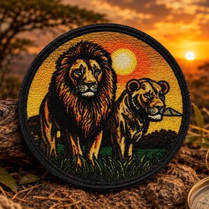 May include: A circular embroidered patch featuring a male lion with a dark mane and a lioness standing in tall green grass. The background depicts a vibrant orange and yellow sunset with a bright sun. The patch has a thick black border.