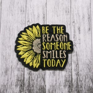 May include: Black embroidered patch with a yellow sunflower and the text "Be the reason someone smiles today."