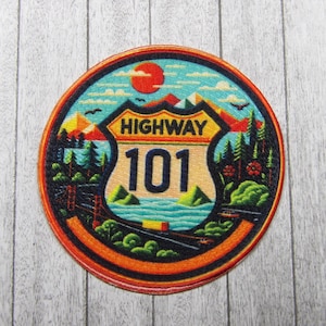 Può includere: Un cerotto rotondo, arancione e blu con un cartello stradale bianco che dice "Highway 101". Il cartello è circondato da un paesaggio colorato con montagne, alberi e un tramonto.