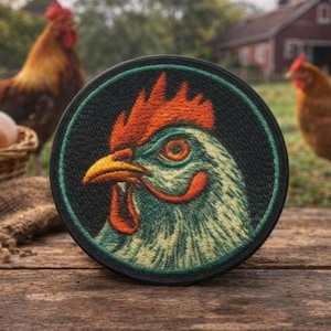 May include: A circular embroidered patch featuring a detailed profile of a chicken head with a bright orange comb and wattle, a yellow beak, and light-colored feathers, all set against a dark background within a thin teal circular border.