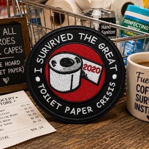 May include: A circular embroidered patch with a black border and a red center. The patch features a white toilet paper roll illustration with the text '2020' on the paper and the words 'I SURVRVED THE GREAT TOILET PAPER CRISIS' in white.
