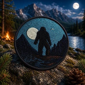 May include: Round embroidered patch featuring a black silhouette of Bigfoot standing before a mountain range, a full moon, and a river. The patch has a blue background and a black border. The patch is set against a natural outdoor background.