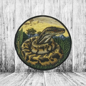 Puede incluir: Un parche redondo bordado con una serpiente marrón y amarilla con ojos rojos, enroscada en un follaje verde sobre un fondo de puesta de sol amarillo y naranja.