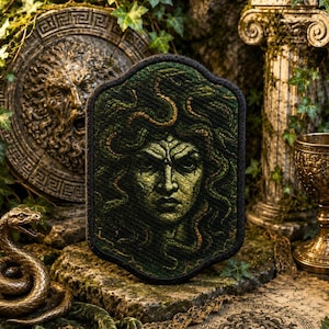 May include: A rectangular patch with rounded corners featuring a dark green and black embroidered portrait of Medusa with snakes for hair. The design is rendered in shades of deep forest green and black thread with a thick black stitched border.